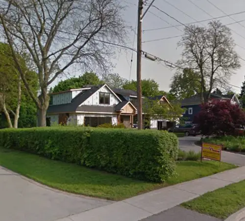 Street view of a two-storey home with a peaked gable behind a mature hedge