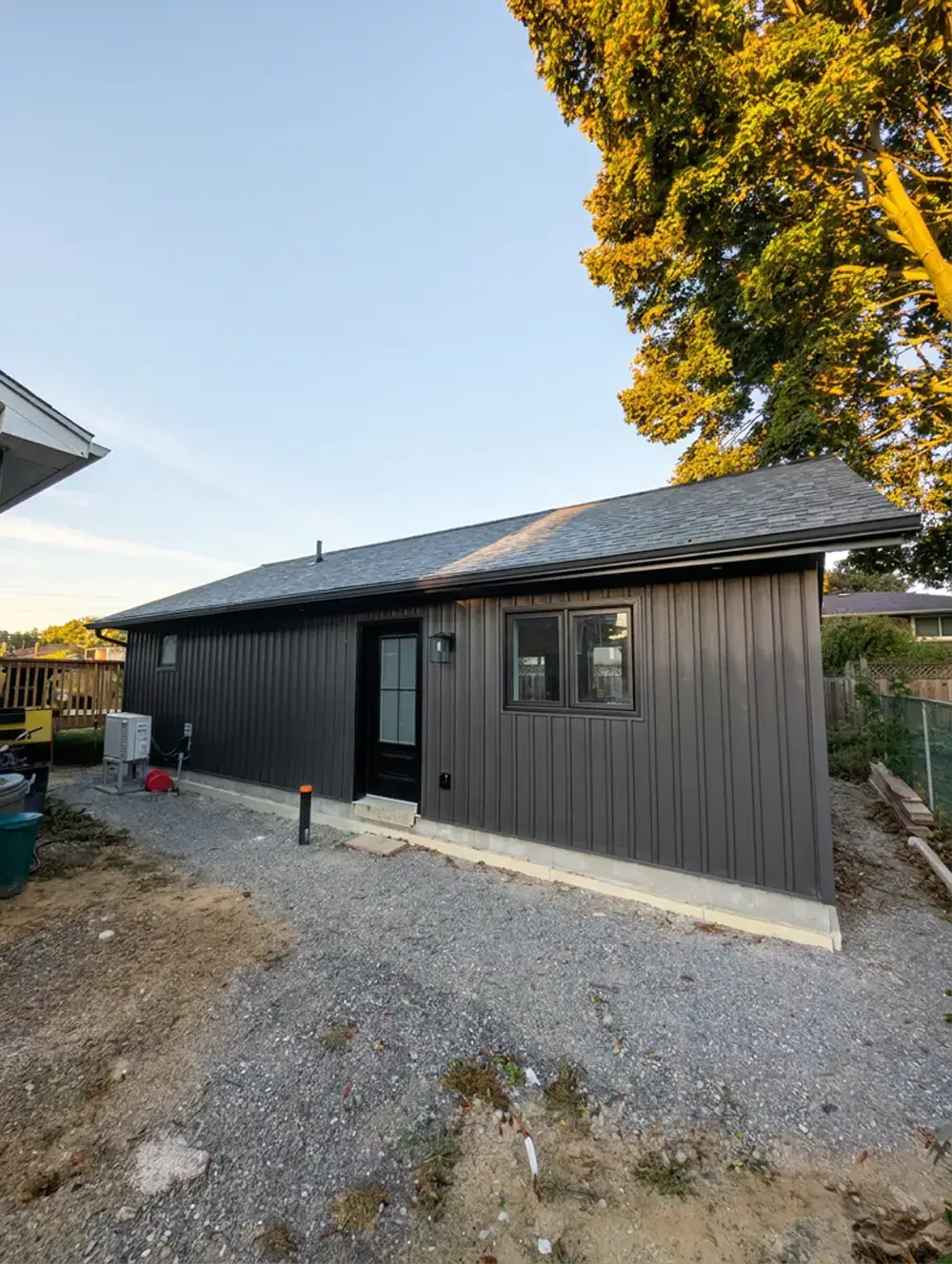Newly built accessory dwelling unit with dark board-and-batten siding, a black entry door and a shingled roof, set at the back of a gravel lot